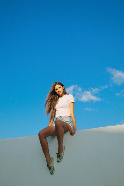 女人穿着白色的背心和蓝色牛仔短裤坐在白墙在湛蓝的天空
